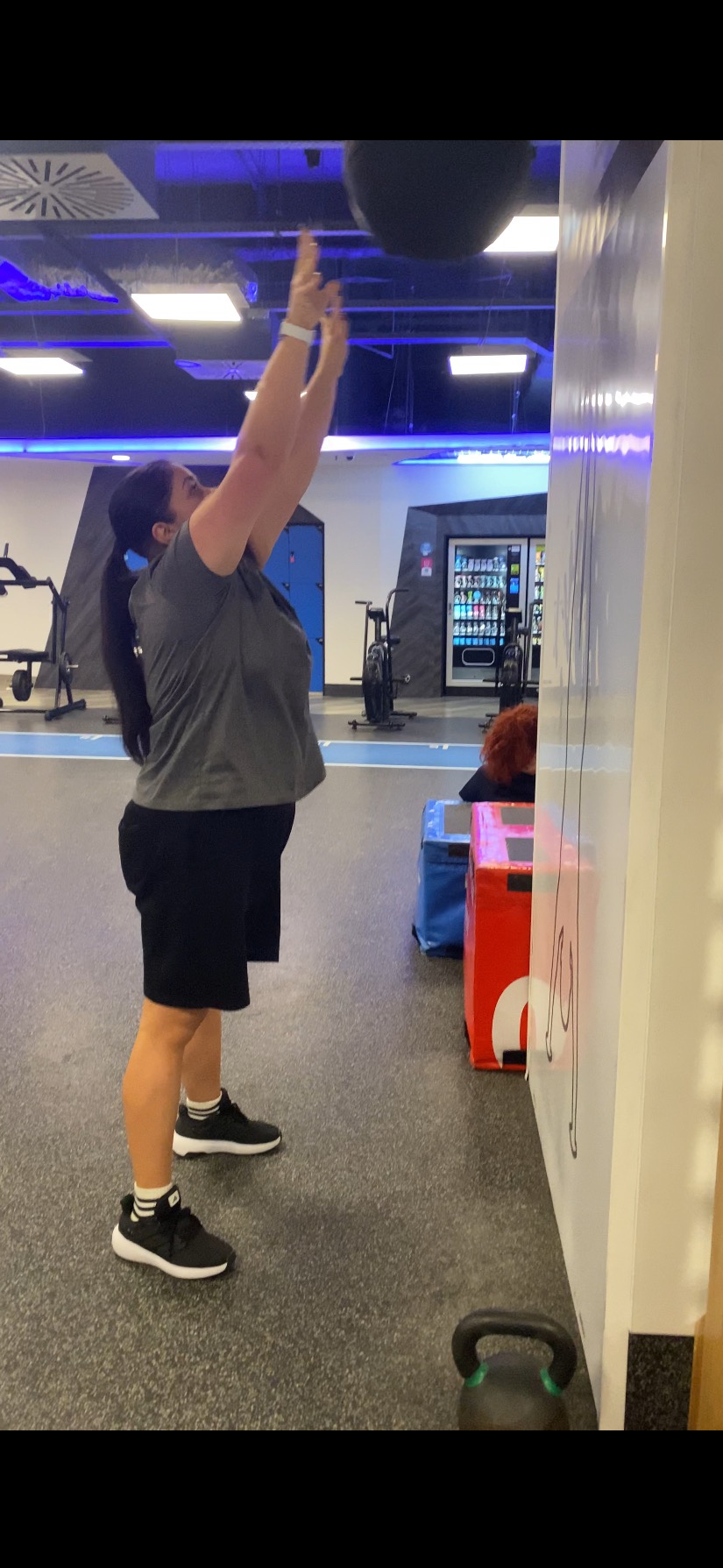 Kobieta w szarym t-shircie ćwiczy z piłką lekarską w nowoczesnej siłowni z niebieskim oświetleniem. W tle automat z napojami i sprzęt do ćwiczeń.