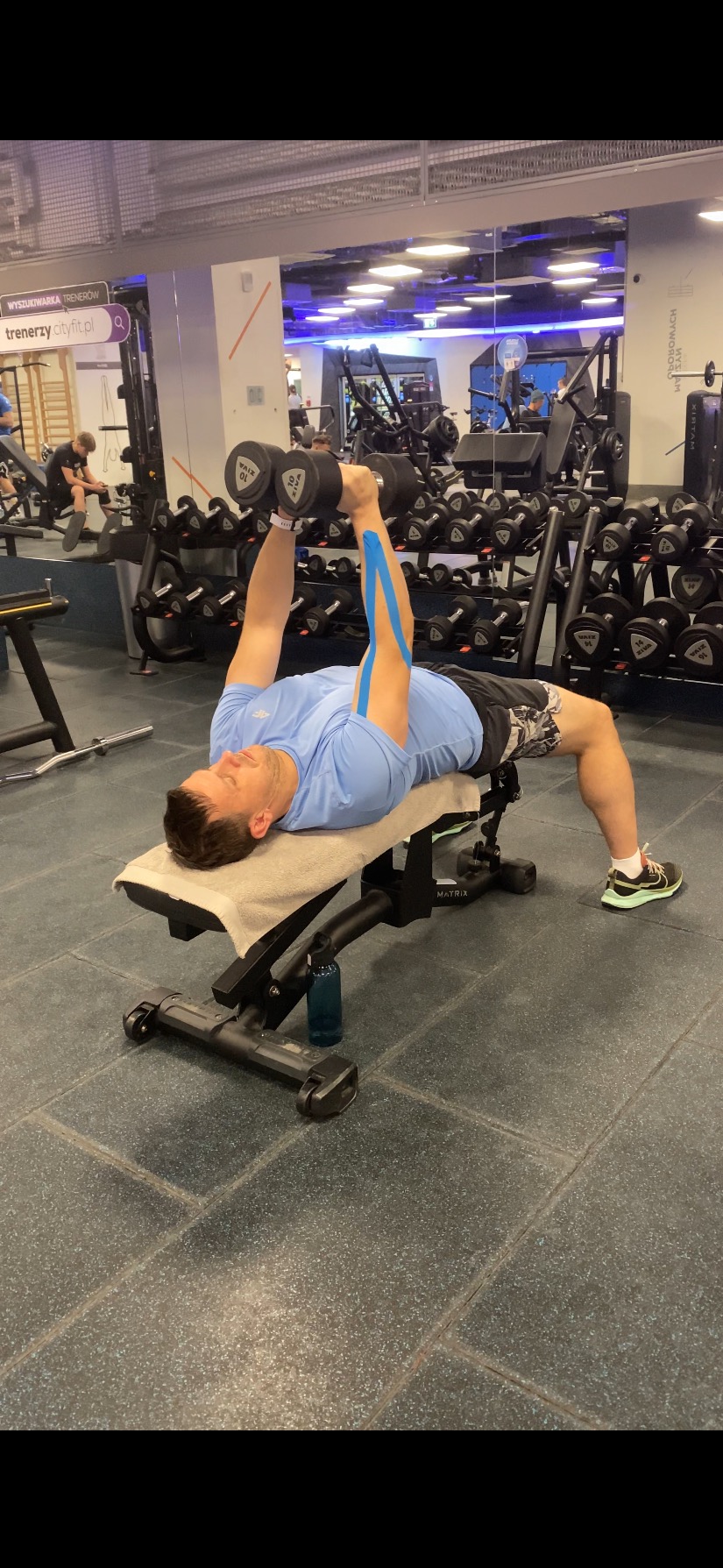 Mężczyzna z niebieskim kinesiotapingiem na ramionach leży na ławce w siłowni, wyciskając hantle. W tle widoczne inne sprzęty i osoby ćwiczące.