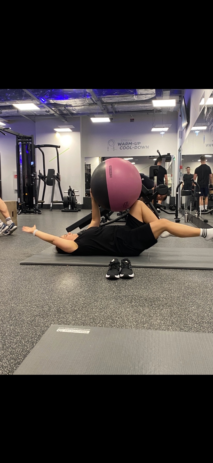 Osoba ćwiczy z piłką fitness na macie w siłowni. Widoczne buty obok maty, lustra i sprzęt do ćwiczeń w tle. Napis 'Strefa Warm-up Cool-down' na ścianie.