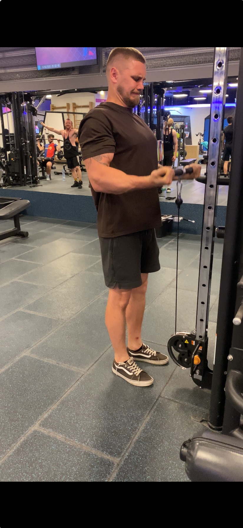 Mężczyzna ćwiczy na maszynie w siłowni, skupiając się na bicepsach. Widoczne tatuaże na ramieniu, sportowe obuwie i inne osoby w tle. Oświetlenie typowe dla siłowni.