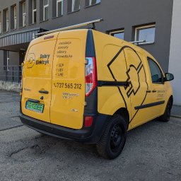Żółty samochód dostawczy z logo 'Dobry Elektryk' i numerem telefonu na tle szarego budynku. Usługi: instalacje, pomiary, odbiory, fotowoltaika. Widoczne oznaczenia SEP, UDT, URE.