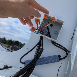 Wojciech Zaczek - Instalacja elektryczna zewnętrzna: ręka łączy przewody w kostkach Wago w puszce z zasilaczem IP67 na tle nieba. Widok z dołu na instalację oświetlenia.