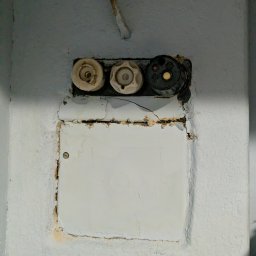 Stara skrzynka z bezpiecznikami topikowymi na pomalowanej ścianie. Widoczne trzy bezpieczniki i biała klapka. Instalacja elektryczna w zaniedbanym stanie.