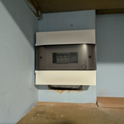 Zamknięta, biała skrzynka elektryczna Legrand z szarą szybką, zamontowana na niebieskiej ścianie w rogu pomieszczenia. Widoczne zabezpieczenia wewnątrz.