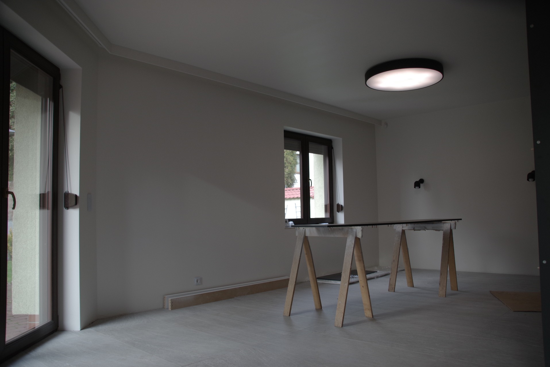 Wnętrze pokoju w trakcie remontu: surowe ściany pomalowane na biało, szare płytki na podłodze, prowizoryczny stół na kozłach, widoczne listwy przypodłogowe w paczkach, okno z widokiem na zieleń...