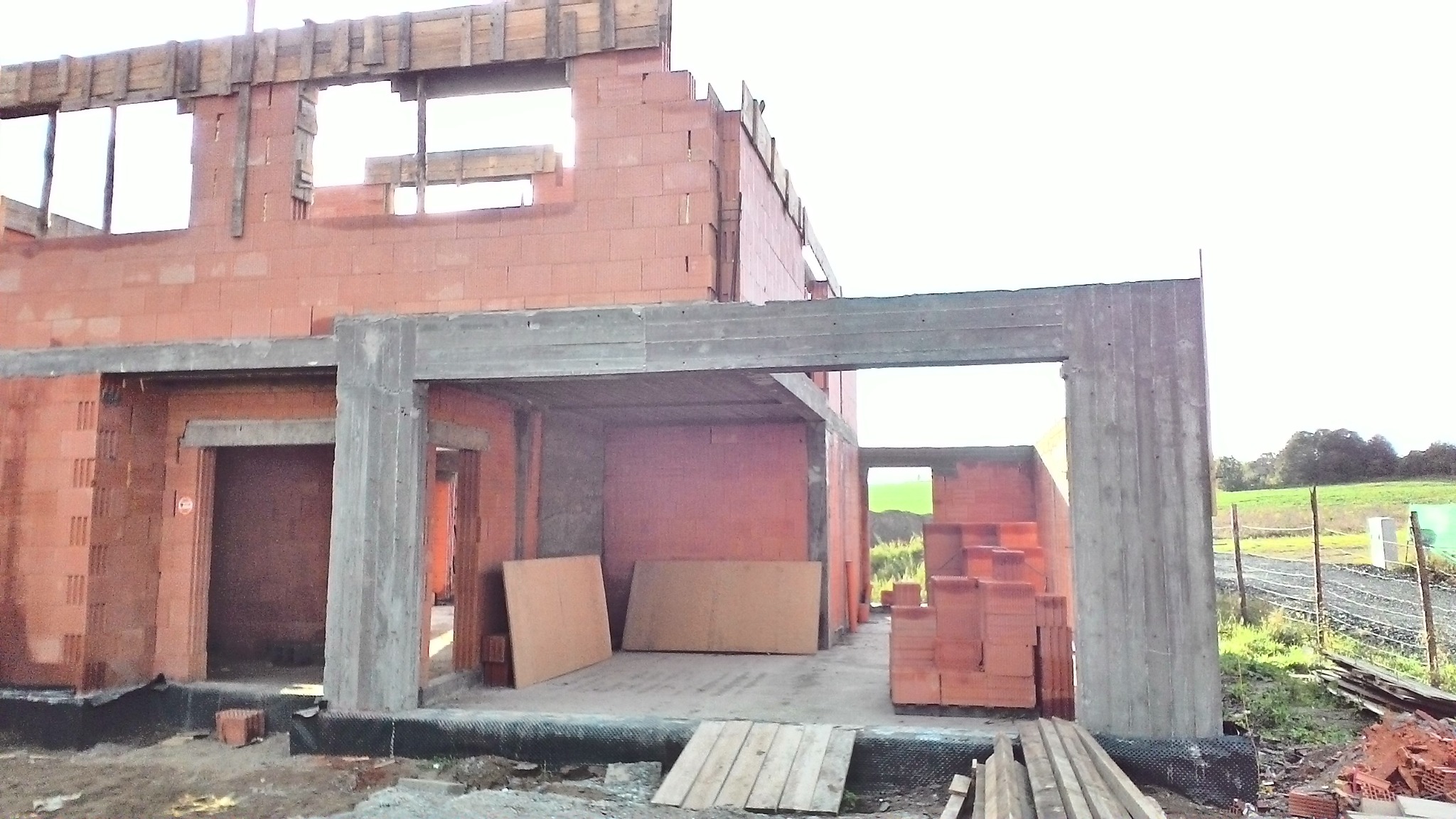 Surowy stan budowy domu jednorodzinnego: widoczne ściany z czerwonej cegły, betonowe filary i drewniane szalunki okienne, z zapasem cegieł i desek na zewnątrz budynku.