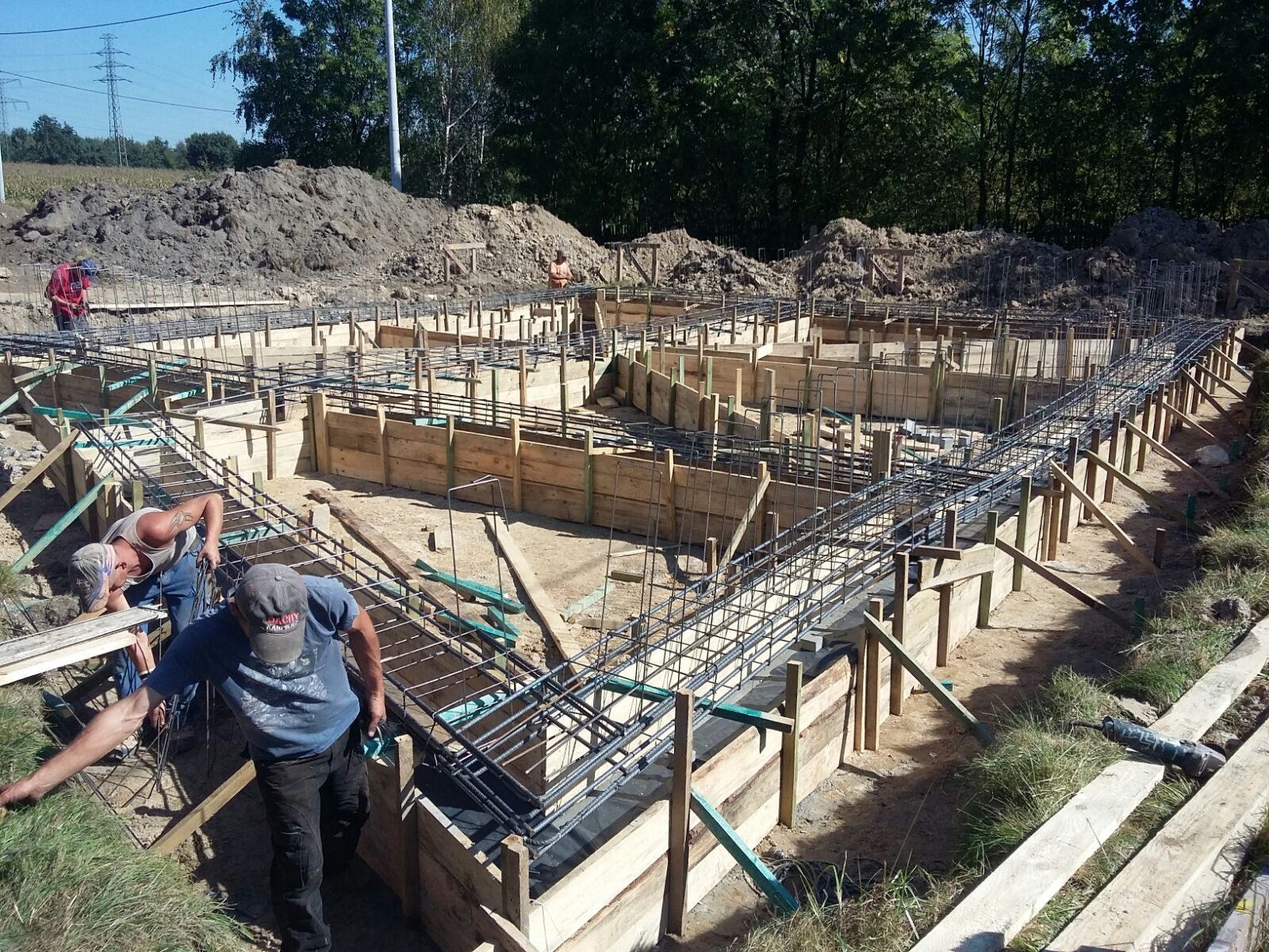 Budowa fundamentów domu jednorodzinnego: szalunki z desek, zbrojenie stalowe, widoczni pracownicy w trakcie montażu, teren budowy z ziemią i drzewami w tle.