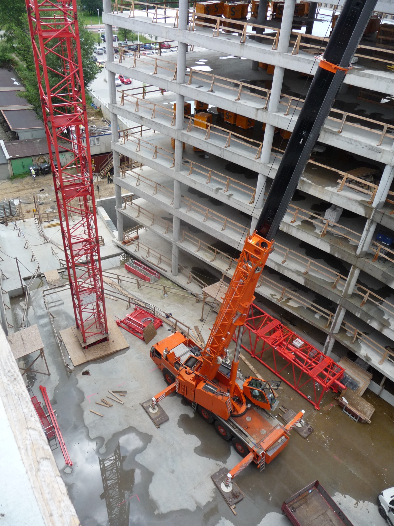 Widok z góry na plac budowy w Katowicach z dwoma dźwigami: pomarańczowym mobilnym i czerwonym wieżowym, przy wznoszącym się żelbetowym budynku w stanie surowym otwartym, po deszczu widać kałuże.