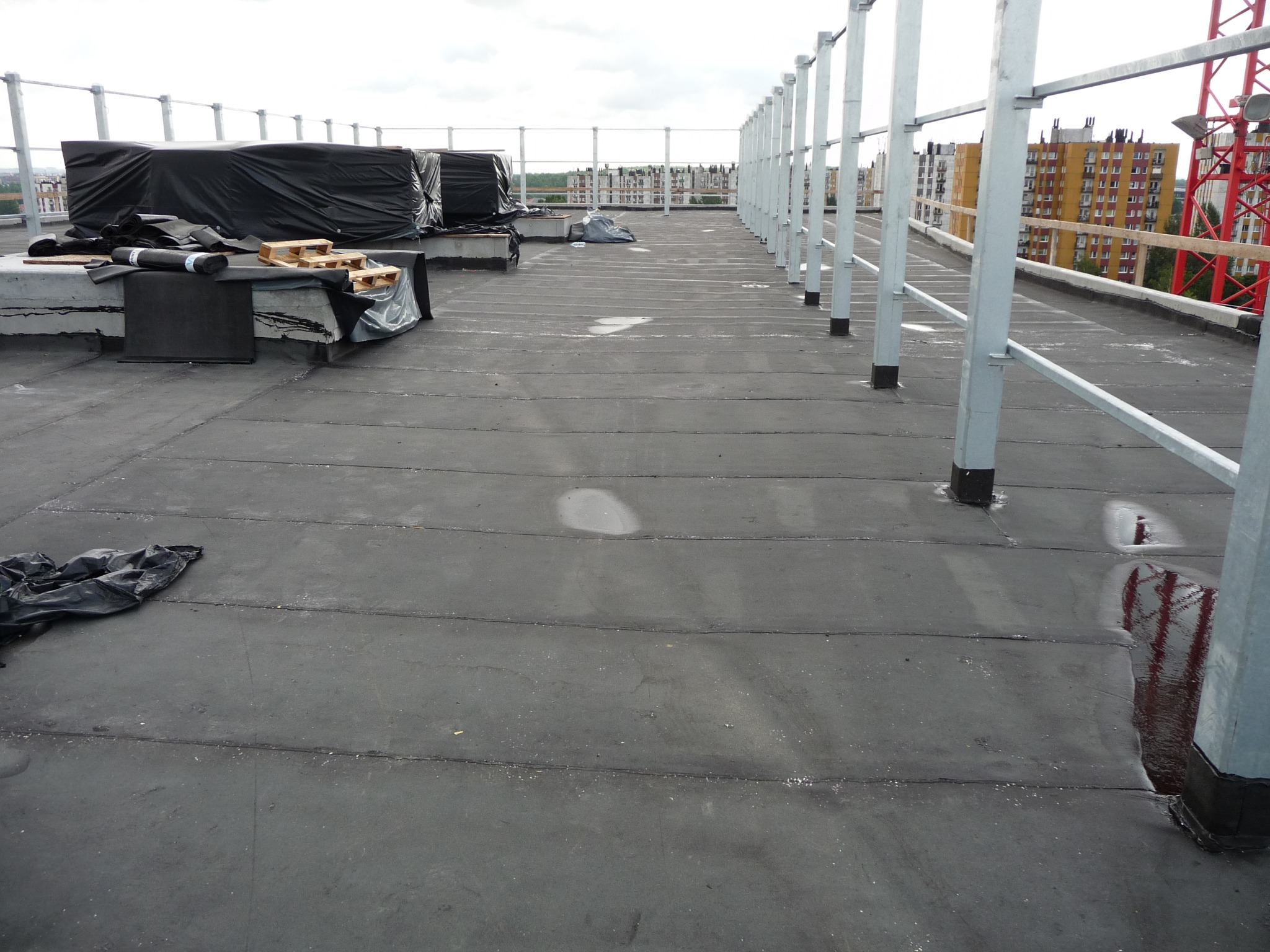 Czarny, płaski dach pokryty papą z widocznymi zgrzewami, z materiałami budowlanymi pod czarną folią i metalowymi barierkami ochronnymi w tle, Katowice.
