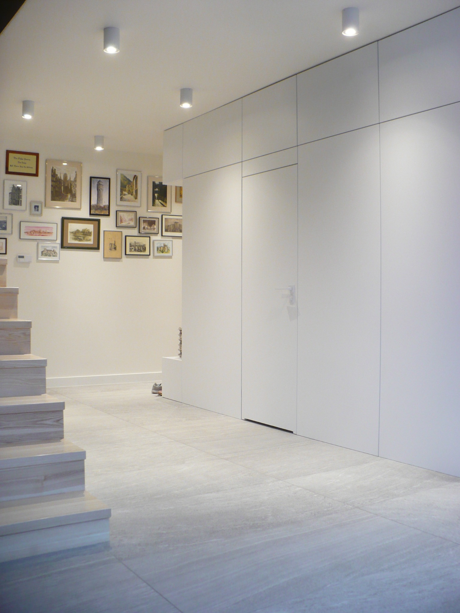 Minimalistyczne wnętrze: białe ściany z wbudowaną szafą, drewniane schody, podłoga z szarych płytek, galeria obrazów na ścianie i punktowe oświetlenie sufitowe.