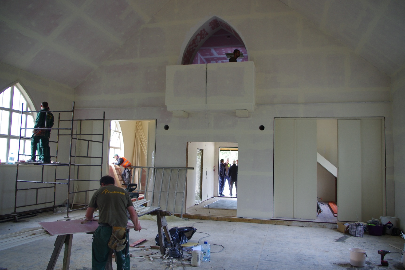 Wnętrze w trakcie wykończenia: pracownicy na rusztowaniach montują płyty gipsowo-kartonowe na ścianach i suficie w przestronnym pomieszczeniu z oknami łukowymi i antresolą, widoczne narzędzia...