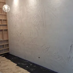 Ściana z dekoracyjną, strukturalną tapetą w odcieniach beżu, zabezpieczona folią malarską na podłodze, widoczne narzędzia malarskie i wiadro z farbą.