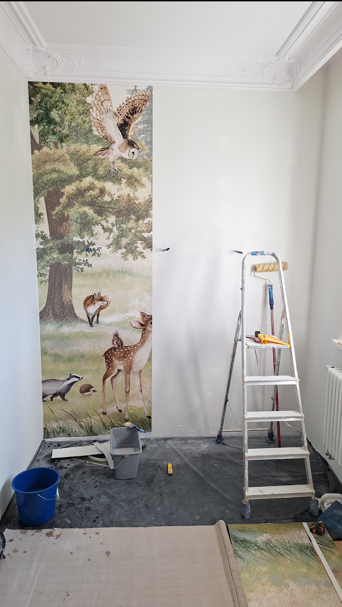 Pokój w trakcie tapetowania: na ścianie przyklejony fragment tapety z motywem leśnym (sowa, jeleń, lis, borsuk, jeż), drabina z narzędziami malarskimi stoi obok, niebieskie wiadro i pojemnik...