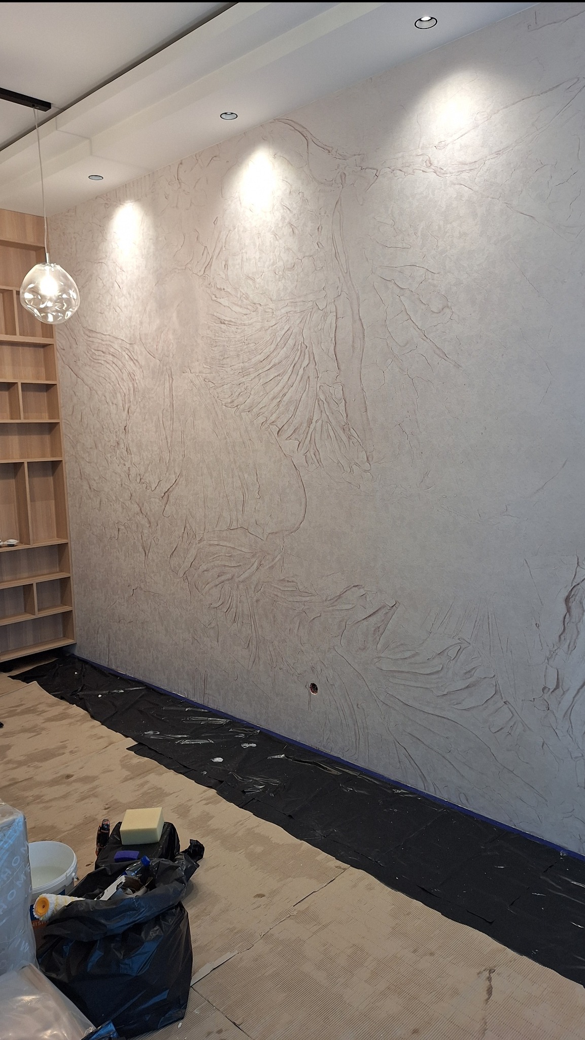 Ściana z dekoracyjną, strukturalną tapetą w odcieniach beżu, zabezpieczona folią malarską na podłodze, widoczne narzędzia malarskie i wiadro z farbą.