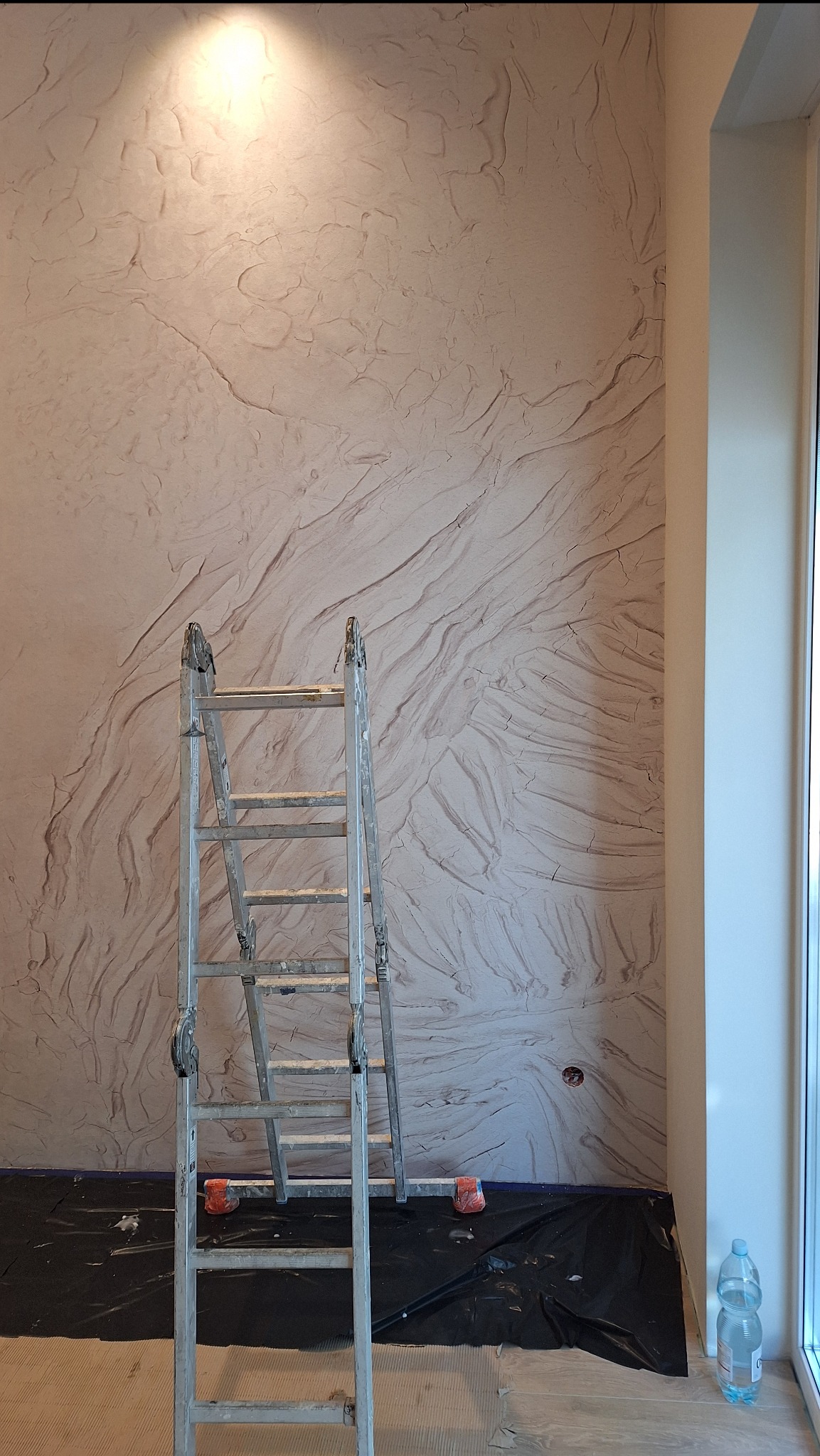Ściana z dekoracyjną fakturą przypominającą naturalny kamień, oświetlona punktowo, z rozstawioną drabiną i folią ochronną na podłodze.