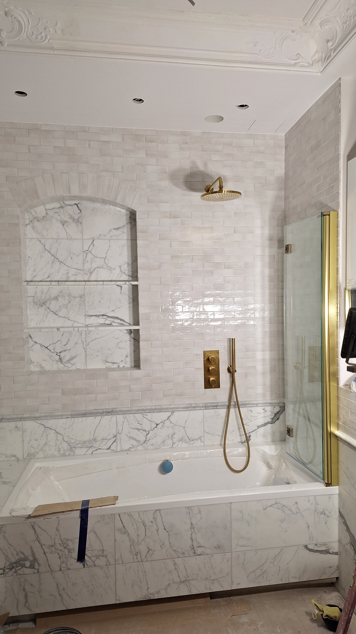 Łazienka w trakcie wykańczania: białe płytki imitujące cegłę na ścianach, obudowa wanny i wnęka z półkami wyłożone płytkami imitującymi marmur, złota armatura i kabina prysznicowa ze złotymi ramami...