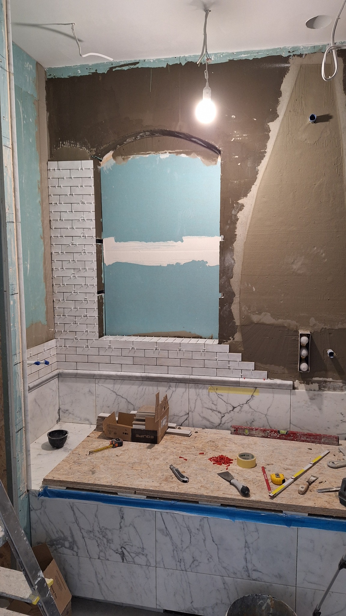 Układanie białych, podłużnych płytek na ścianie w łazience, widoczne krzyżyki dystansowe, narzędzia na zabezpieczonej folią wannie, ściana w trakcie przygotowania.