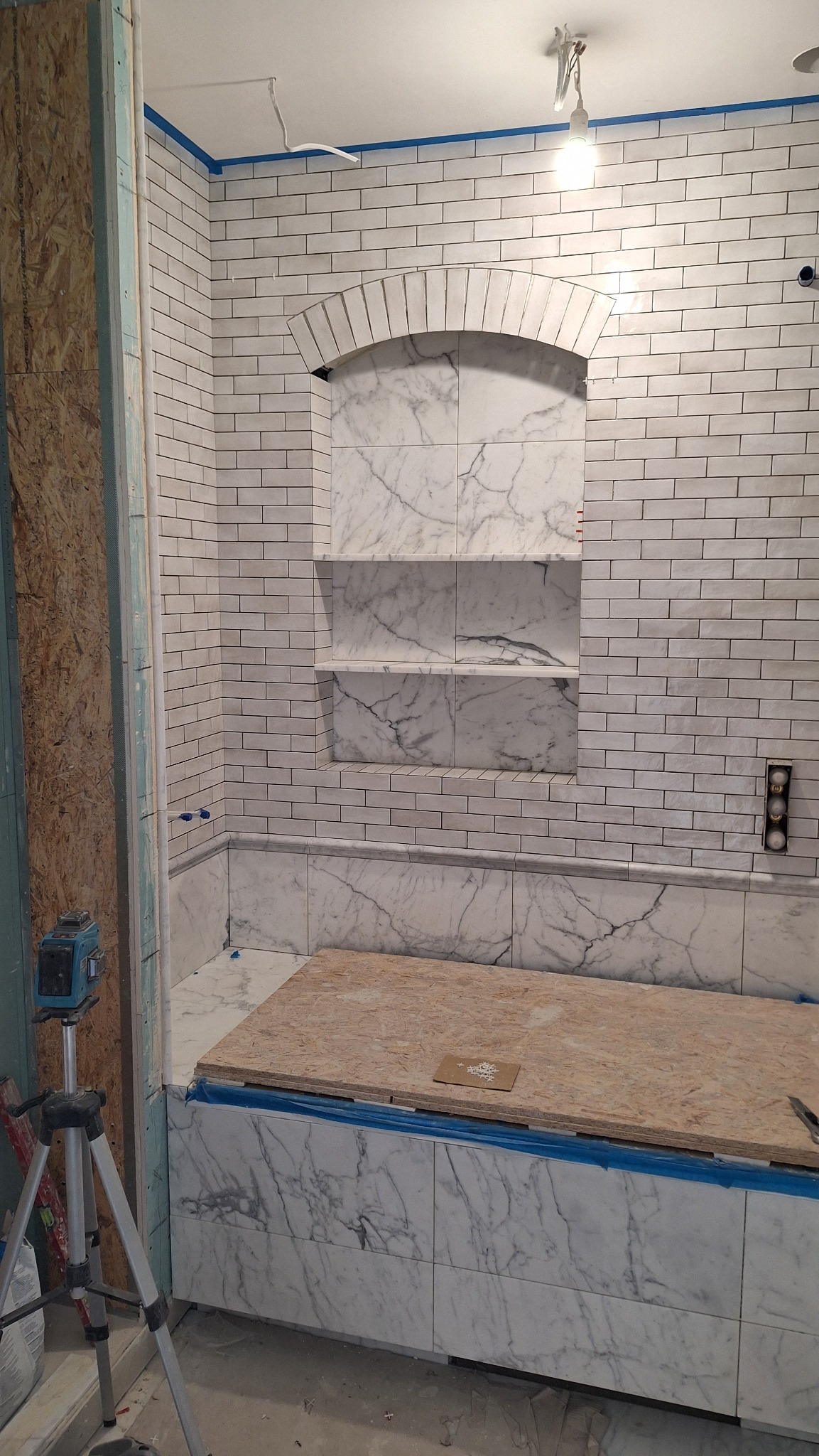 Wnętrze łazienki w trakcie remontu: białe, prostokątne płytki na ścianie, wnęka z marmurowymi półkami zwieńczona łukiem, obudowa wanny wyłożona marmurem, zabezpieczona niebieską taśmą, widoczny...