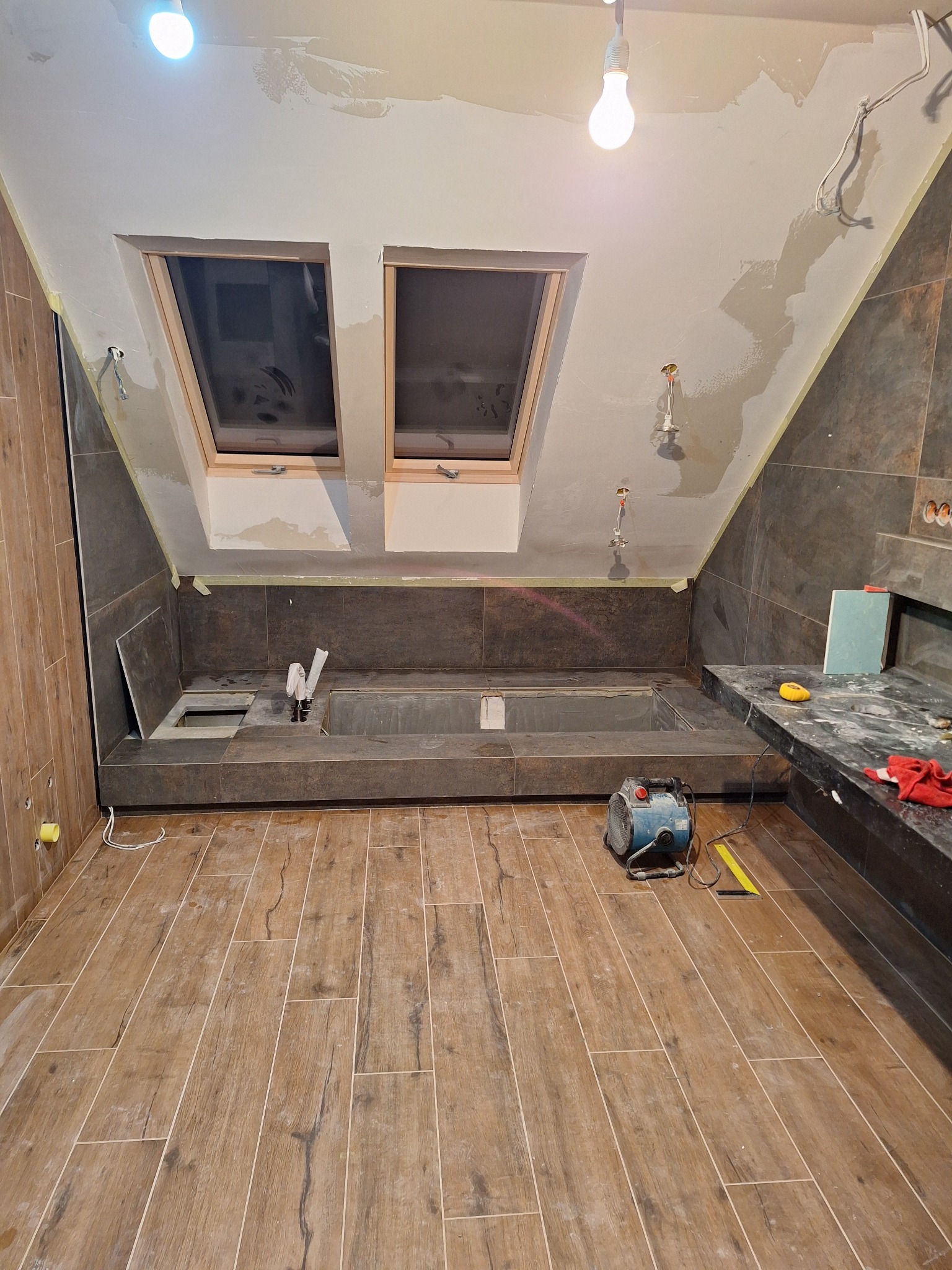 Adaptacja poddasza: montaż brązowych płytek imitujących kamień na ścianach i podłodze, widoczne okna dachowe, przygotowane miejsce na brodzik oraz nagrzewnica budowlana.