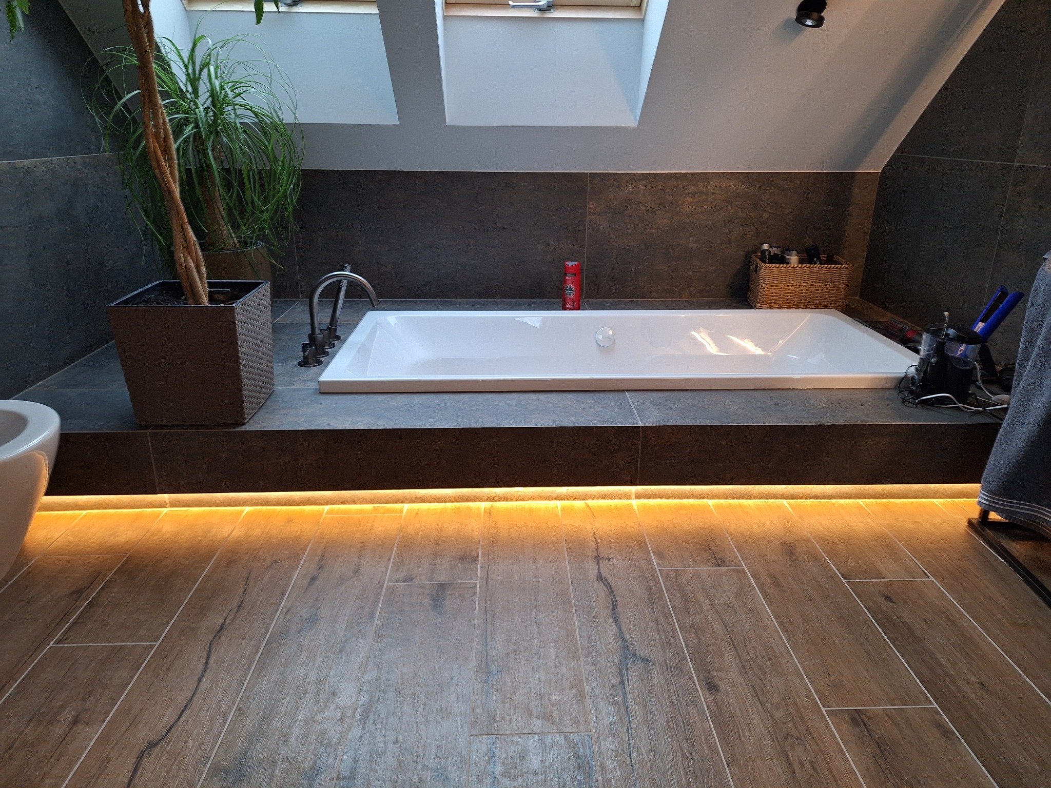 Nowoczesna łazienka na poddaszu z wanną, drewnopodobnymi płytkami podłogowymi z podświetleniem LED, szarymi płytkami ściennymi i rośliną w donicy.