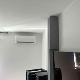 BartHome Bartłomiej Chrapek - Biała klimatyzacja ścienna w minimalistycznym wnętrzu z szarą kolumną i sprzętem audio. Nowoczesny design, subtelne oświetlenie LED.