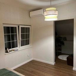 BartHome Bartłomiej Chrapek - Biała klimatyzacja ścienna w jasnym pokoju z drewnianą podłogą, widoczna nad oknem. W tle lampa wisząca i otwarte drzwi do kolejnego pomieszczenia.