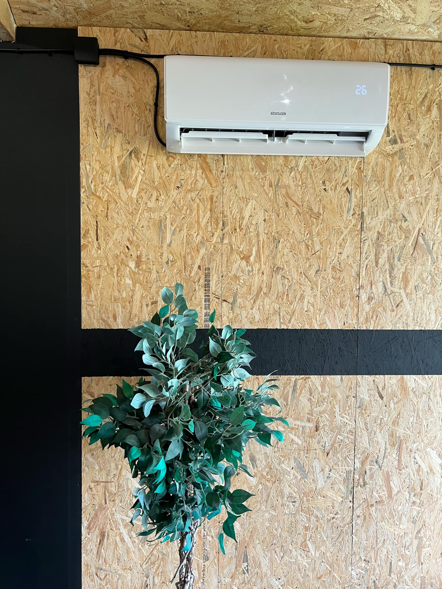 Biały klimatyzator Sinclair zamontowany na ścianie z płyty OSB, z widocznymi kablami i dekoracyjną rośliną w rogu.