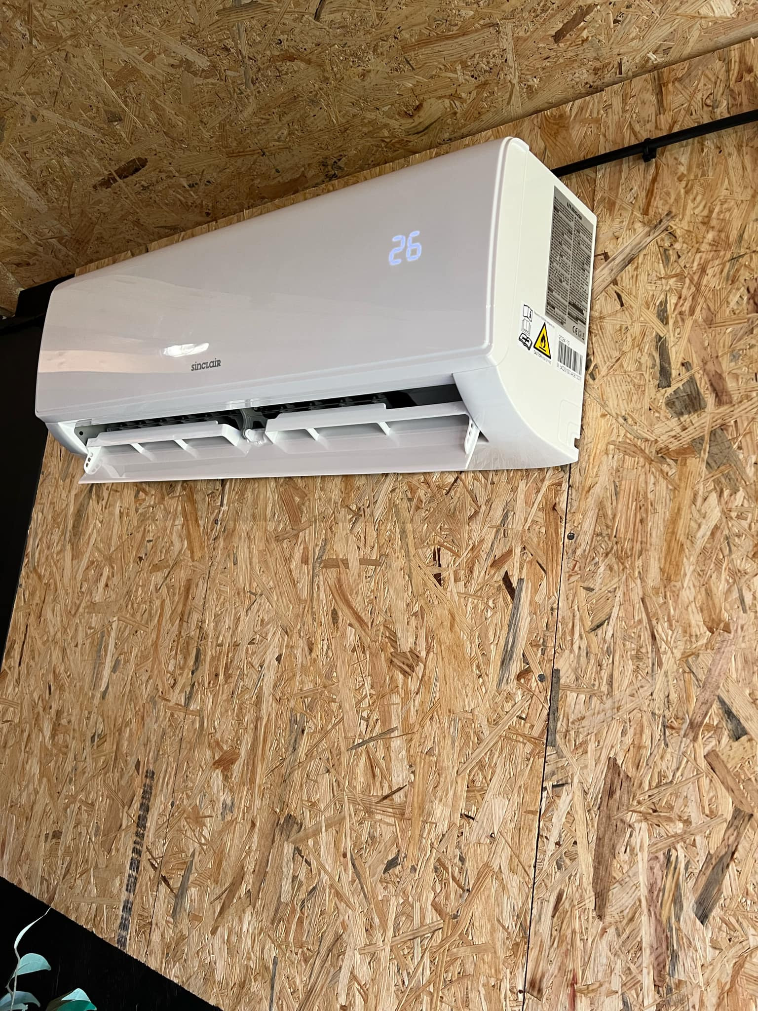 Biała klimatyzacja Sinclair zamontowana na ścianie z płyty OSB, wyświetlająca temperaturę 26 stopni.