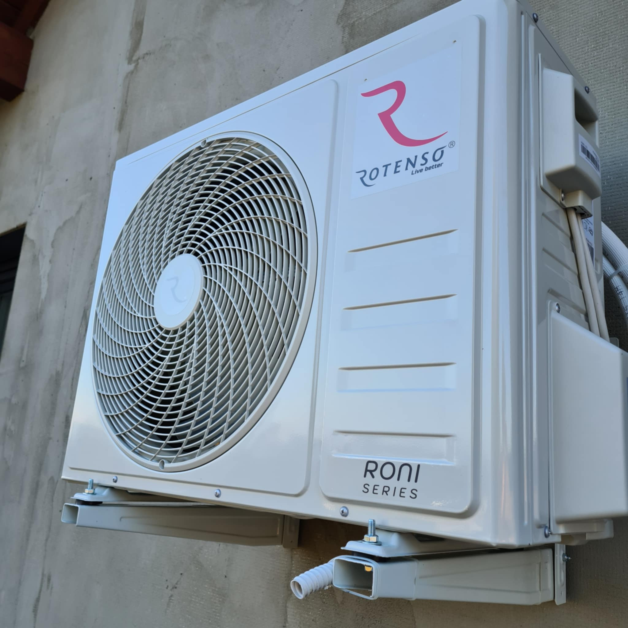 Biała jednostka zewnętrzna klimatyzatora marki Rotenso, model RONI Series, zamontowana na szarej ścianie budynku, widoczne przewody i elementy montażowe.