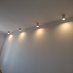 Usługi Elektryczne Szymon Zaganiaczyk - Rząd cylindrycznych, białych reflektorów punktowych LED zamontowanych na suficie w jasnym pomieszczeniu, oświetlających białą ścianę. Widoczne okno z lewej strony i fragment nagrzewnicy Master...