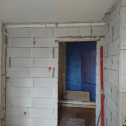 Wnętrze w trakcie remontu z widocznymi przewodami elektrycznymi prowadzonymi po ścianach z betonu komórkowego, zabezpieczone przejście folią i poziomica oparta o ścianę.