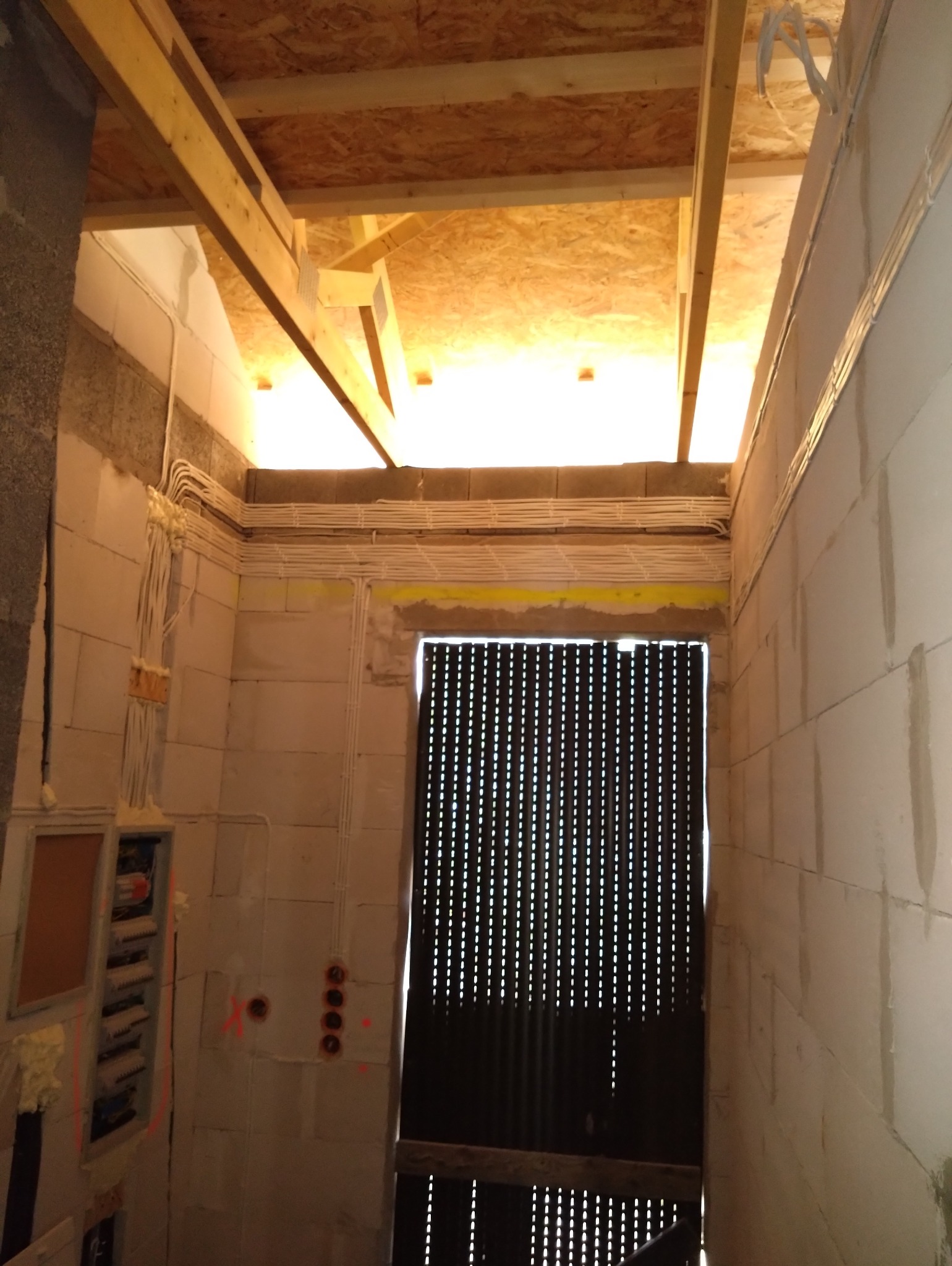 Wnętrze budynku w stanie surowym: widoczne kable elektryczne prowadzone po ścianach z bloczków, skrzynka rozdzielcza, otwory na gniazdka, drewniana konstrukcja dachu i metalowe drzwi z licznymi...