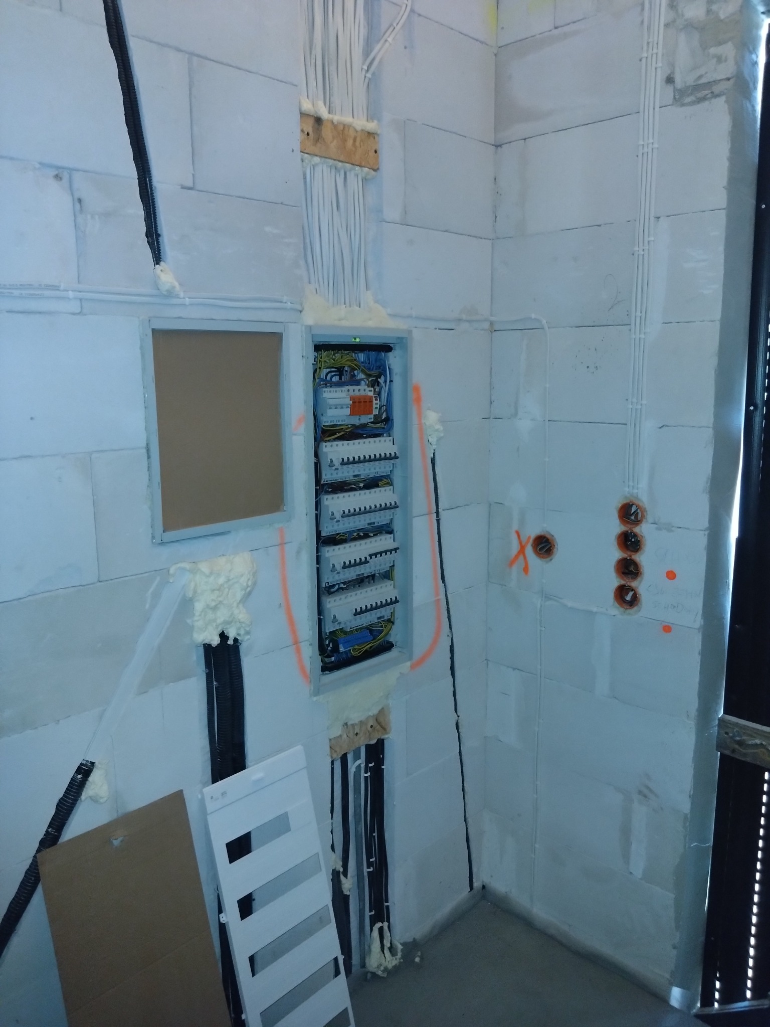 Wnętrze w trakcie prac elektrycznych: rozdzielnica z zamontowanymi bezpiecznikami, kable prowadzone w ścianie z betonu komórkowego, puszki elektryczne z pomarańczowymi oznaczeniami, widoczne pianka...