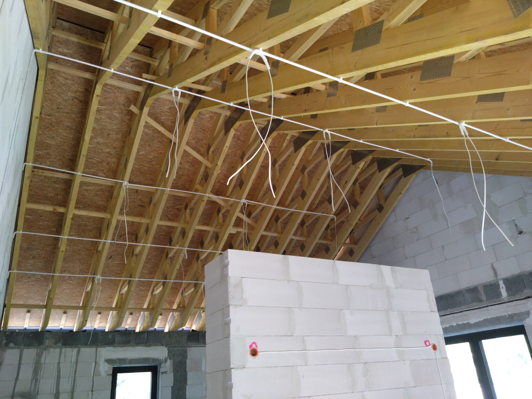 Wnętrze w trakcie budowy z widoczną drewnianą konstrukcją dachu, białymi ścianami z betonu komórkowego i prowizorycznie poprowadzonymi przewodami elektrycznymi w białych rurkach mocowanych...
