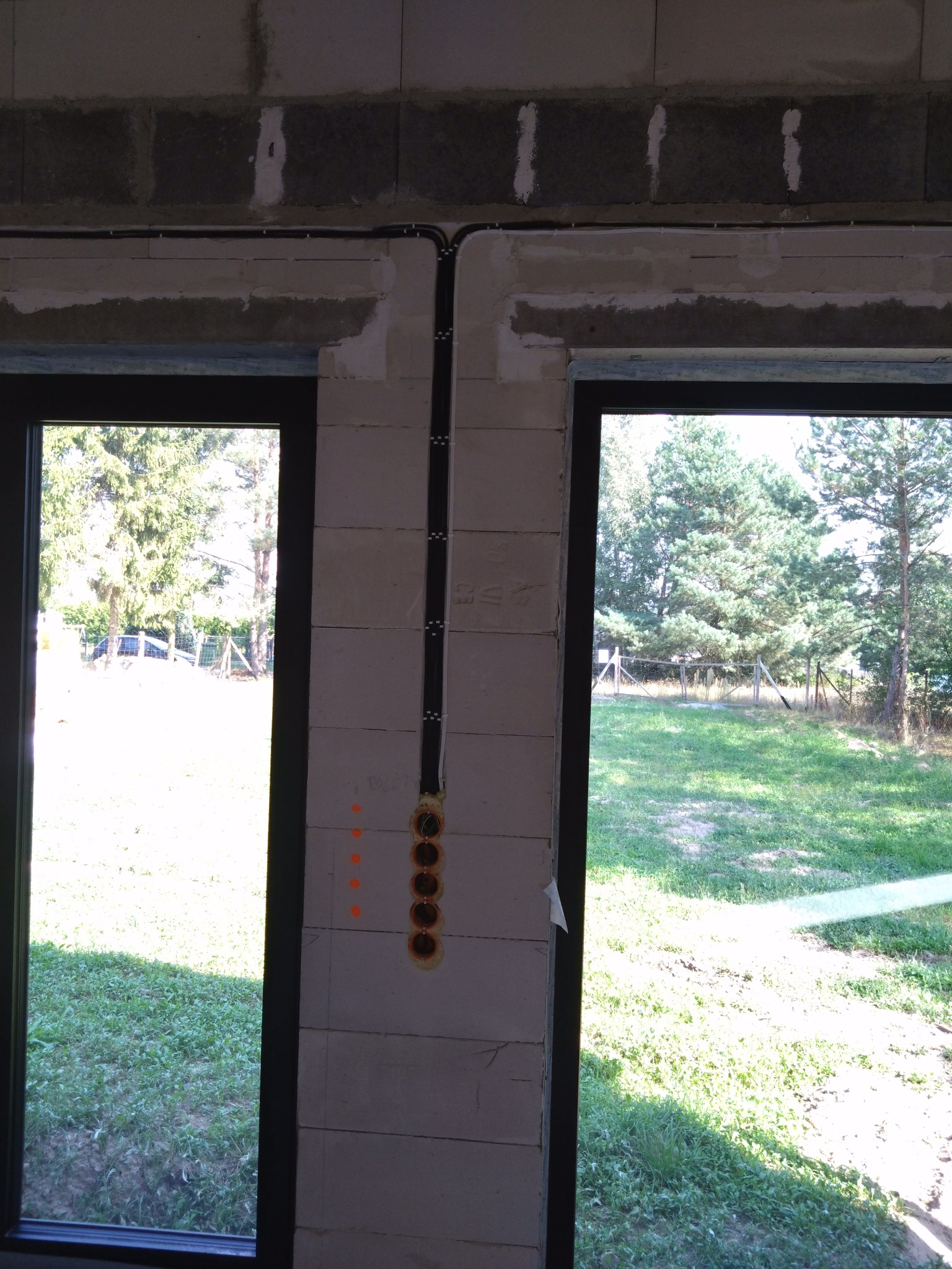 Wewnętrzna ściana z bloczków, z pionowo poprowadzonym kablem elektrycznym w peszlu i puszkami pod gniazdka, widok przez okno na zewnątrz.