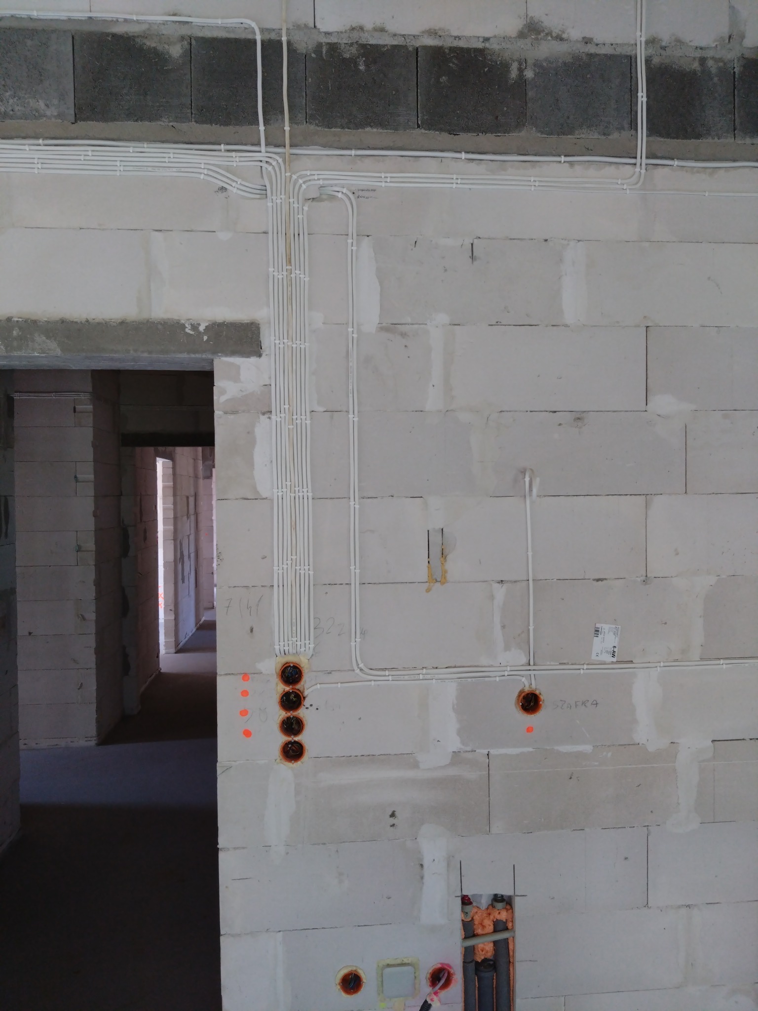 Surowa ściana z betonu komórkowego w trakcie budowy, z widocznymi peszlami elektrycznymi prowadzonymi po powierzchni i wpuszczanymi w otwory, przygotowane pod gniazdka i włączniki, pomarańczowe...