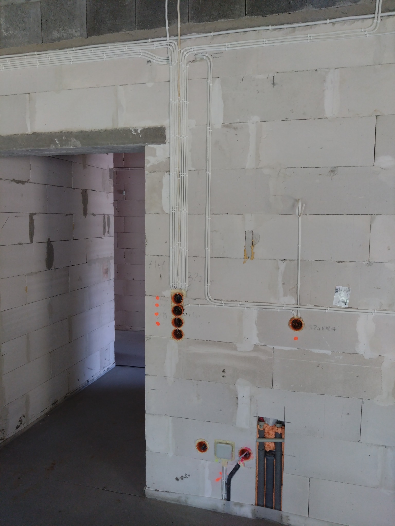 Ściana z betonu komórkowego z widocznymi, nowymi instalacjami elektrycznymi w białych rurkach PCV, prowadzącymi pionowo i poziomo, z wywierconymi otworami pod gniazdka i puszkami instalacyjnymi...