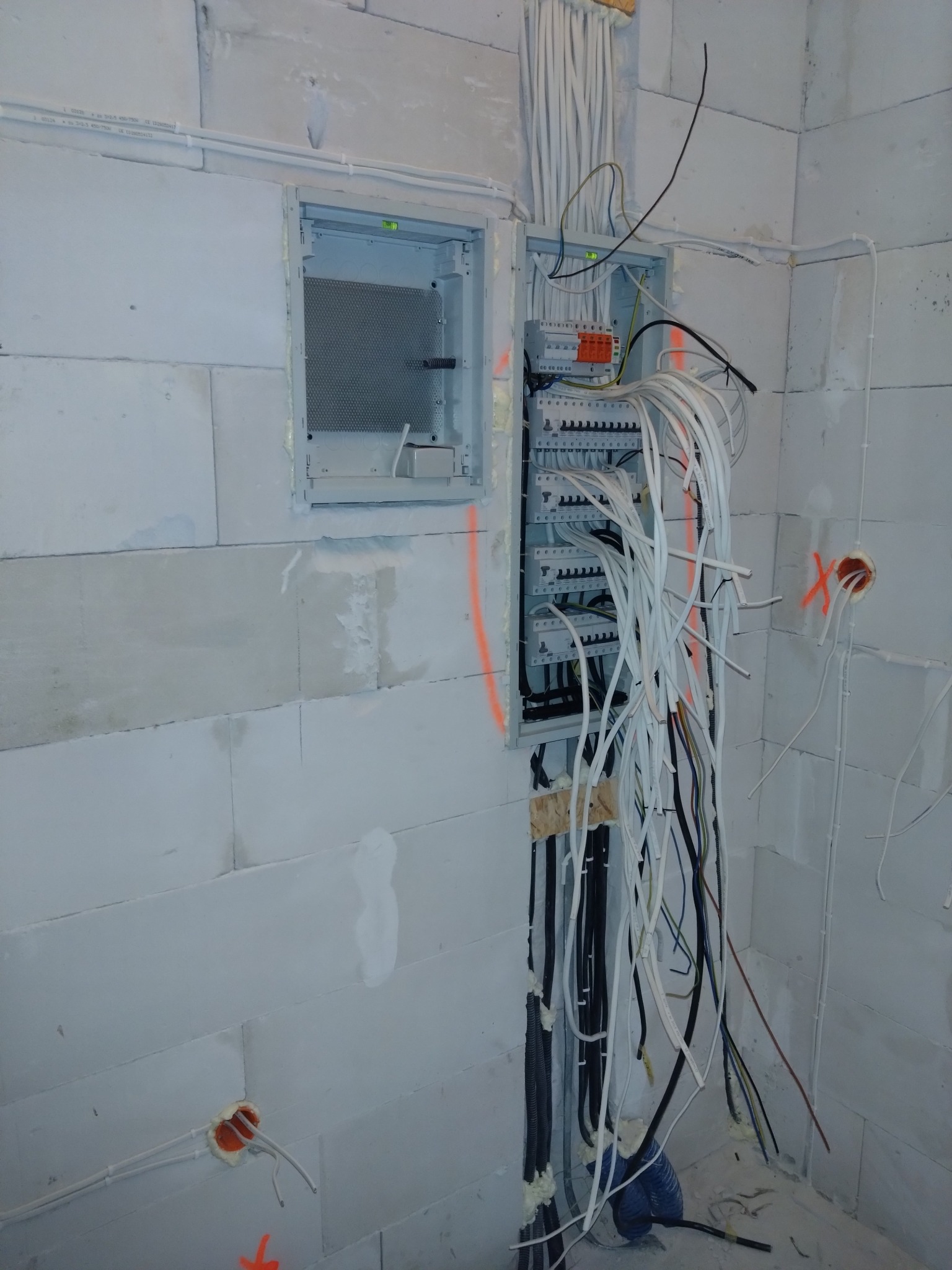 Rozdzielnia elektryczna w surowym stanie, z licznymi kablami wychodzącymi na tle ściany z betonu komórkowego. Widoczne otwory pod gniazdka z wystającymi przewodami, oznaczone pomarańczowym sprayem.