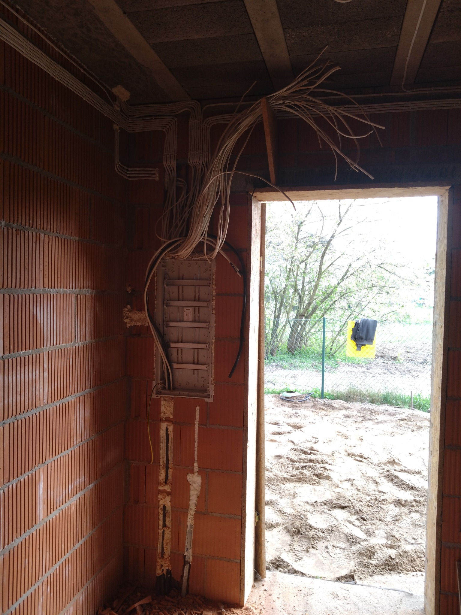 Wnętrze w trakcie prac wykończeniowych z widoczną, częściowo zamontowaną skrzynką elektryczną na ceglanej ścianie, wystającymi przewodami i bruzdami pod kable wypełnionymi gipsem; w tle widok...