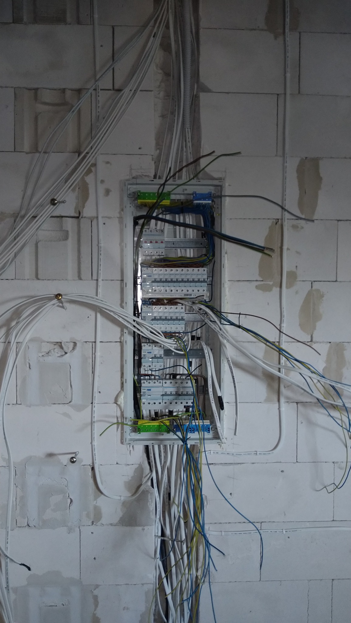 Rozdzielnica elektryczna z licznymi przewodami w ścianie z białych bloczków, widoczne zabezpieczenia i złącza, przewody w kolorach białym, niebieskim i zielonym.