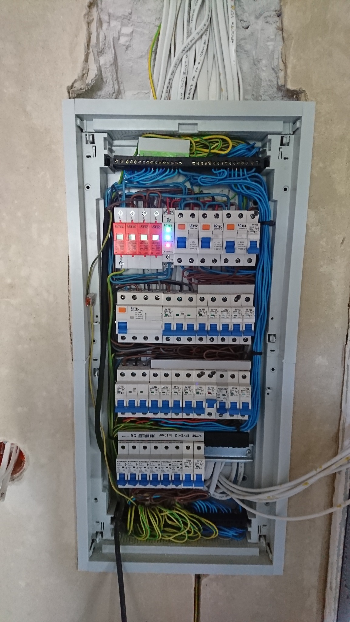 Rozdzielnica elektryczna w szarej obudowie, widoczne zabezpieczenia różnicowoprądowe, bezpieczniki automatyczne, przewody w różnych kolorach oraz listwa zaciskowa. Jeden z ograniczników przepięć...