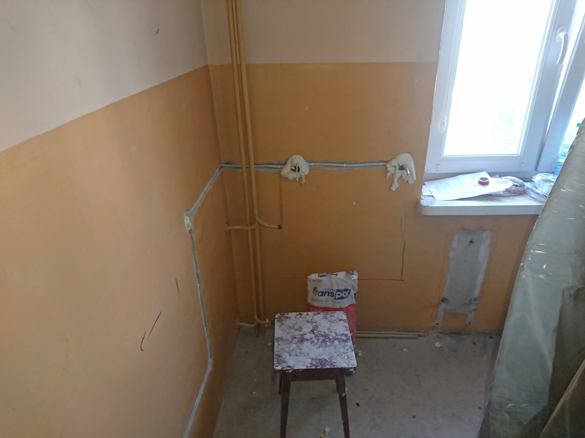 Wągrowiec: Przygotowanie do instalacji elektrycznej w pomieszczeniu z widocznymi przewodami zabezpieczonymi pianką montażową, rurami grzewczymi i workiem z zaprawą murarską stojącym za stołkiem.