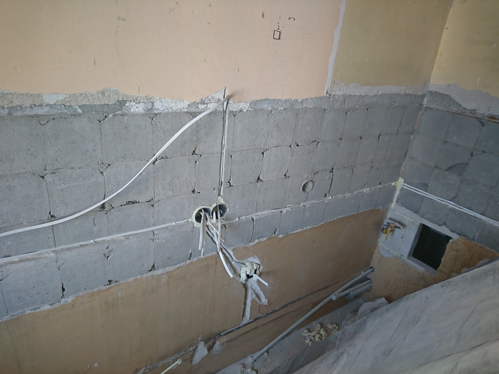 Surowy stan ściany z betonu komórkowego z widocznymi przewodami elektrycznymi wychodzącymi z otworów, częściowo zabezpieczonymi pianką montażową, oraz fragmentem instalacji hydraulicznej w tle.