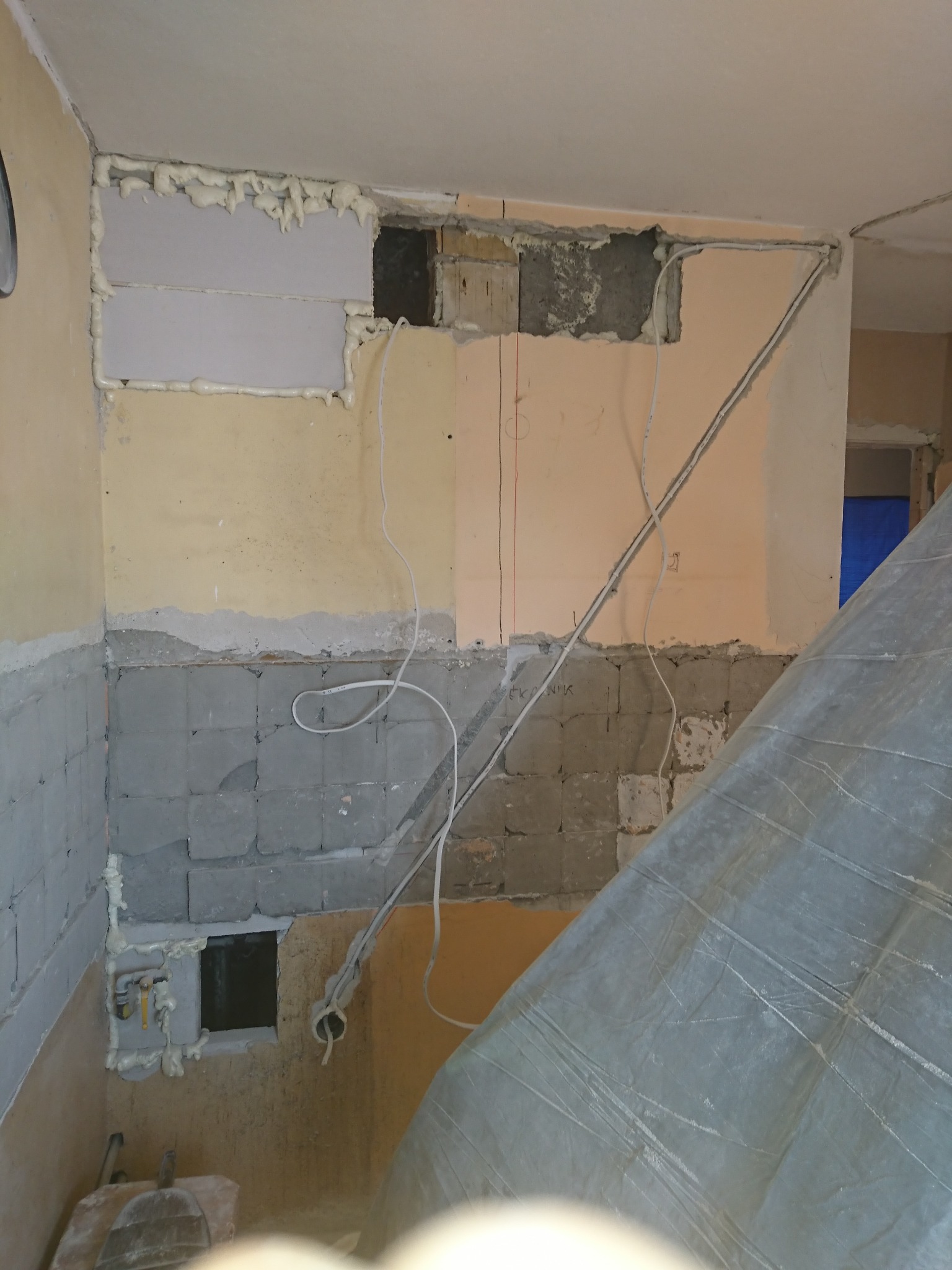 Ściana w trakcie remontu z widocznymi przewodami elektrycznymi, częściowo zakryta folią ochronną, z fragmentami odsłoniętej instalacji i pianką montażową wokół otworu w ścianie.