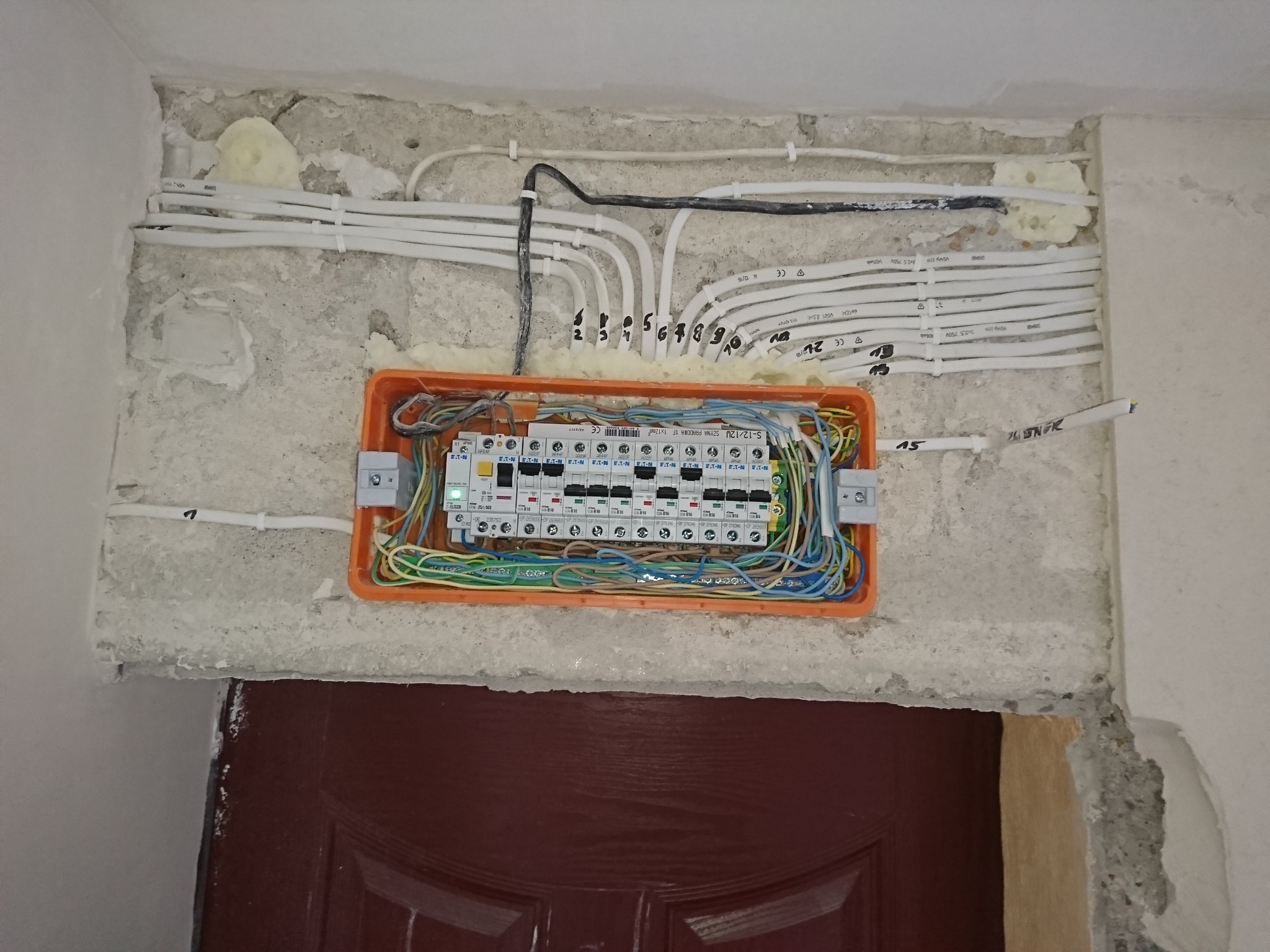 Otwarta pomarańczowa puszka elektryczna z bezpiecznikami i podłączonymi przewodami w ścianie nad framugą drzwi, widoczne numery na przewodach i pianka montażowa wokół.