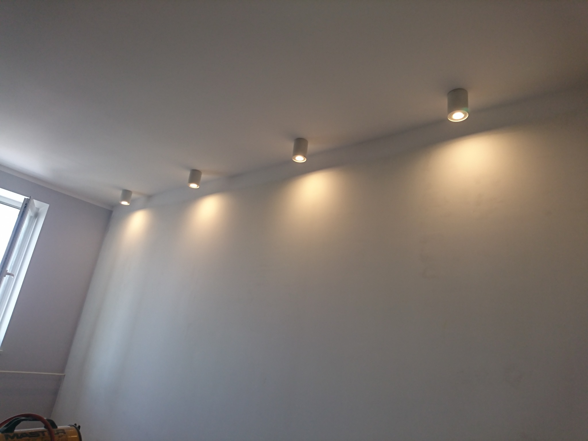 Rząd cylindrycznych, białych reflektorów punktowych LED zamontowanych na suficie w jasnym pomieszczeniu, oświetlających białą ścianę. Widoczne okno z lewej strony i fragment nagrzewnicy Master...