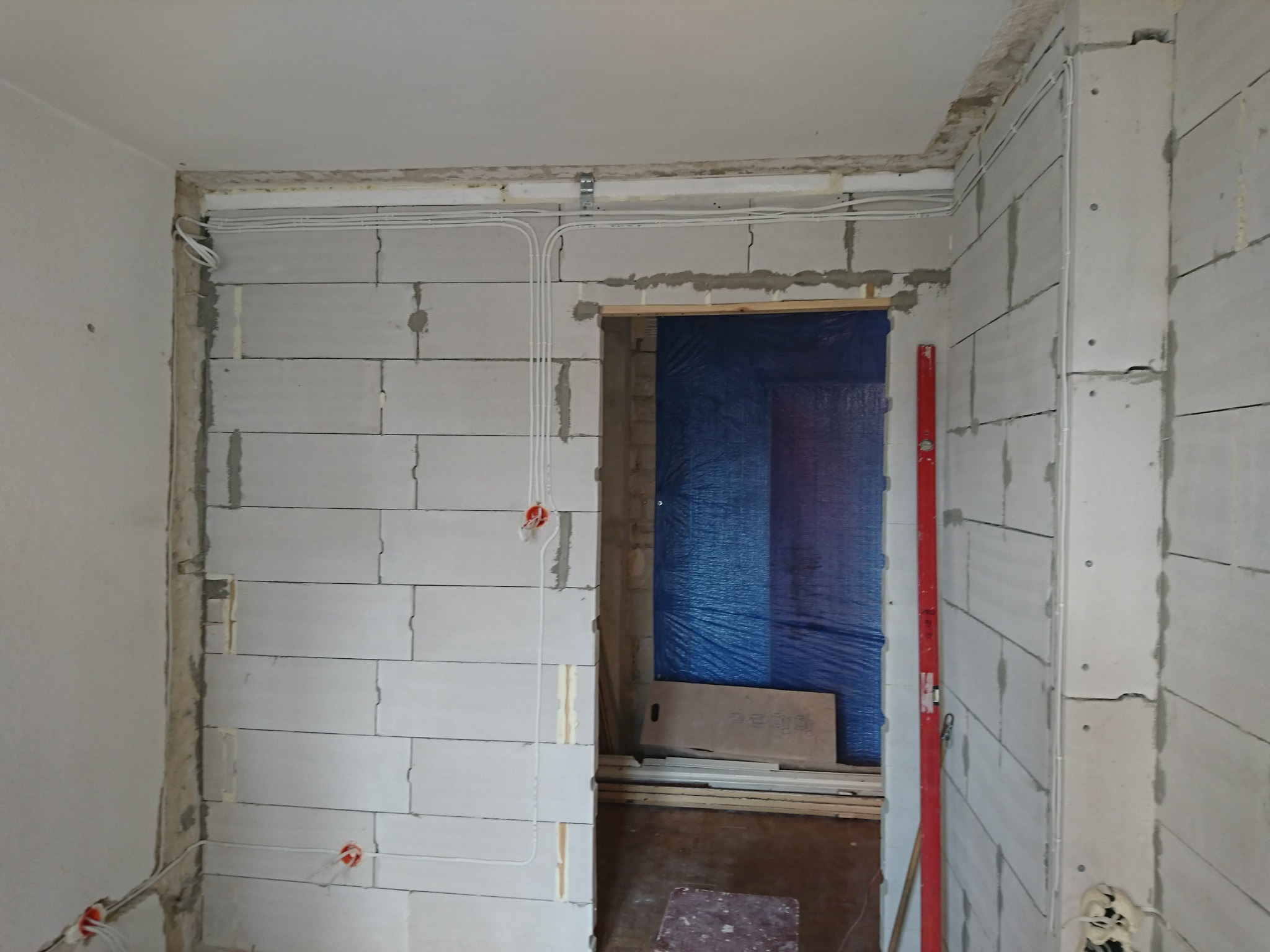 Wnętrze w trakcie remontu z widocznymi przewodami elektrycznymi prowadzonymi po ścianach z betonu komórkowego, zabezpieczone przejście folią i poziomica oparta o ścianę.