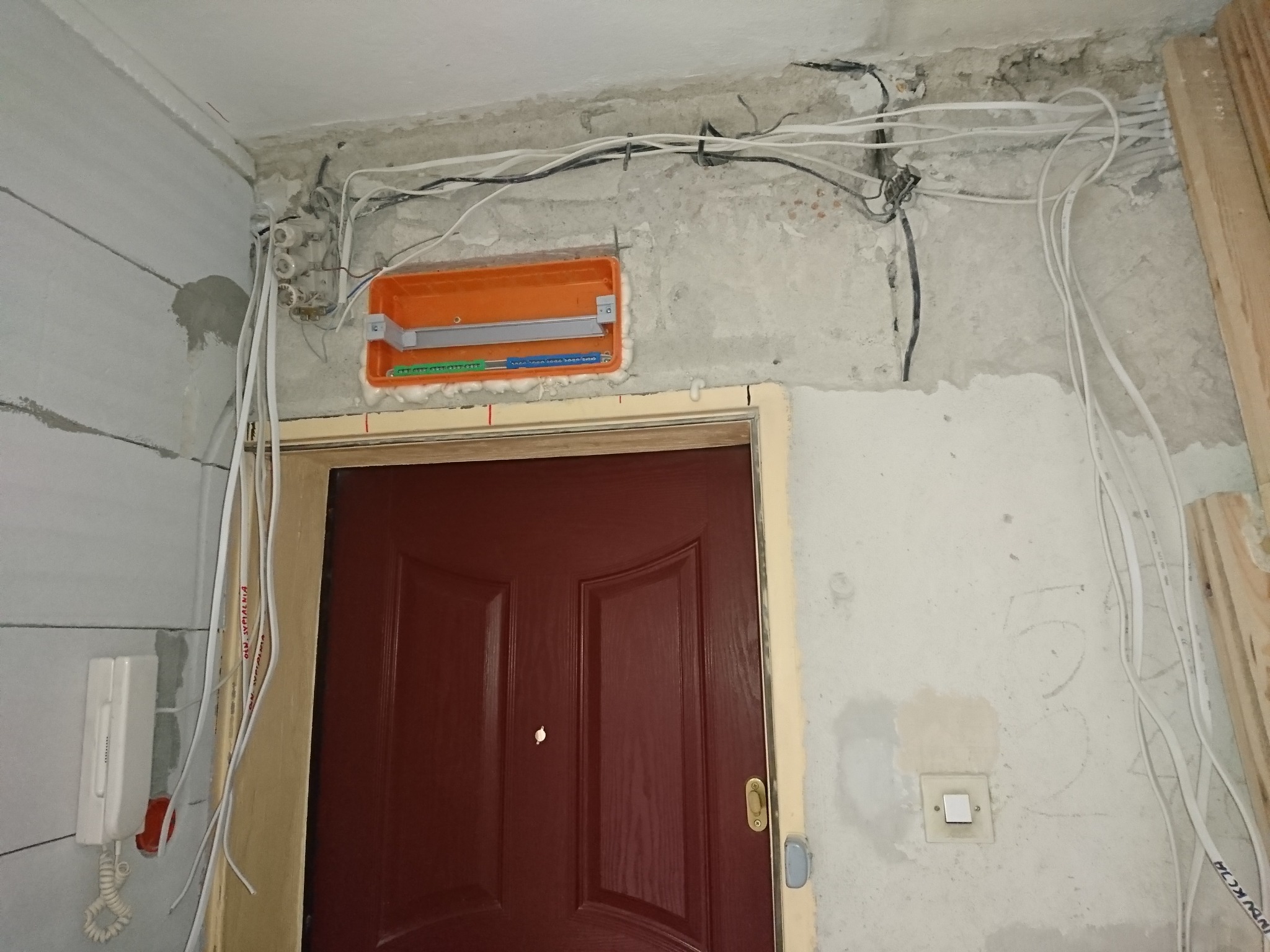 Instalacja elektryczna w trakcie montażu nad drzwiami wejściowymi: widoczna pomarańczowa puszka instalacyjna, białe kable prowadzone po ścianie, stary bezpiecznik topikowy i domofon po lewej stronie