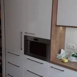 Nowoczesna, biała zabudowa kuchenna z piekarnikiem i szufladami w Czchowie, wykończona drewnopodobnym dekorem, z widocznymi akcesoriami kuchennymi na blacie.