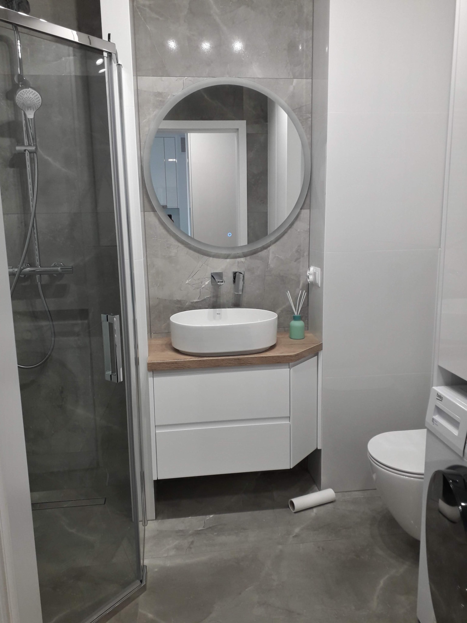 Nowoczesna łazienka z szarą marmurową płytką, okrągłym podświetlanym lustrem nad białą szafką z drewnianym blatem i owalną umywalką, kabina prysznicowa ze szklanymi drzwiami.